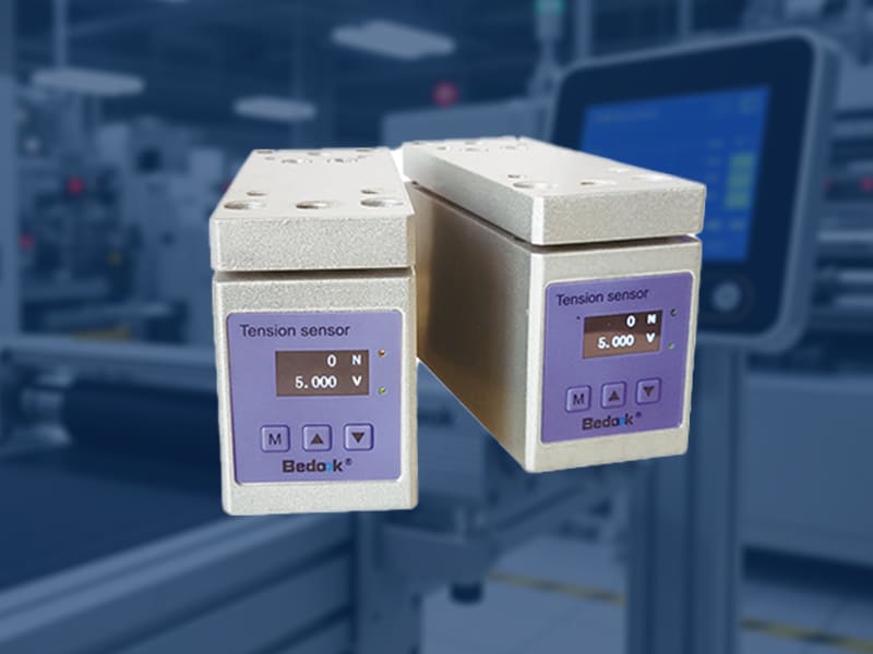 A Bedook TSRI series pillow block web tension sensor installed on a printing press material transfer line, with a display device showing real-time tension data beside it
